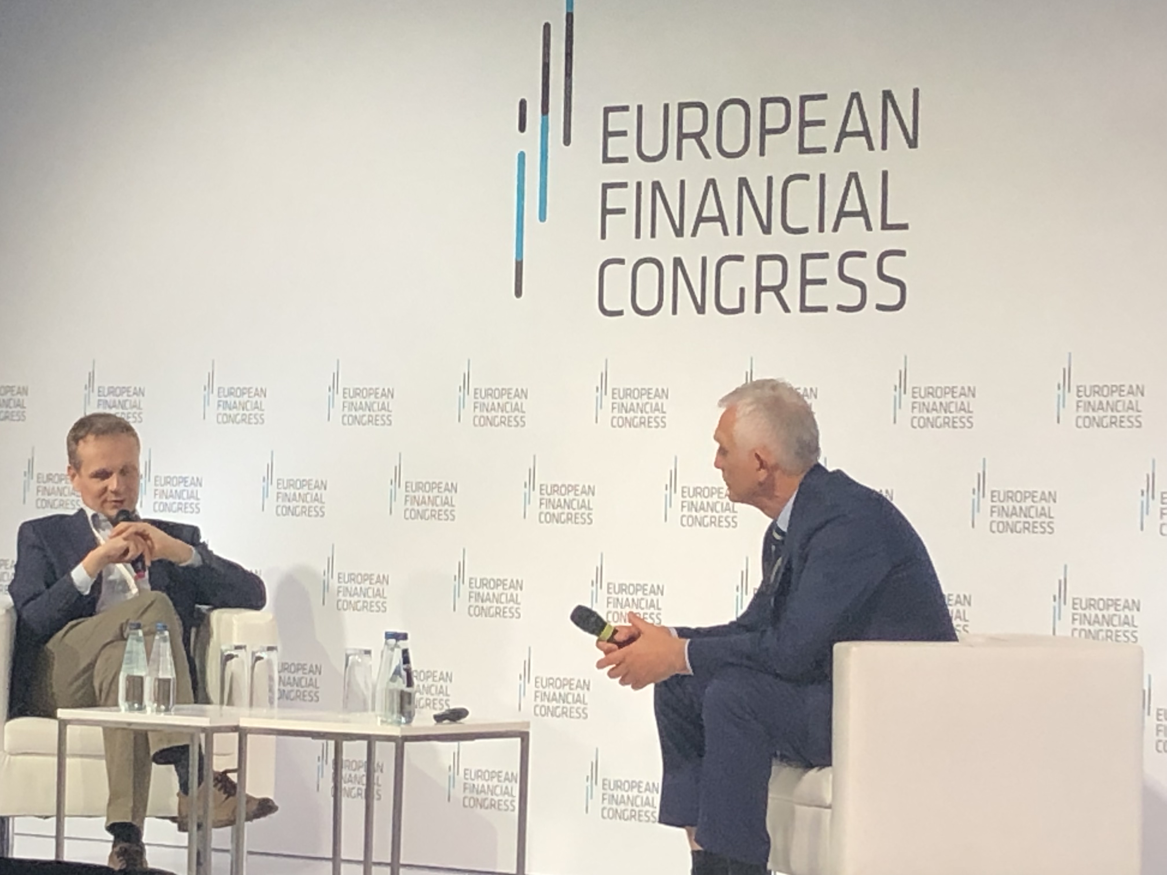 dwóch mężczyzn siedzi na panelu dyskusyjnym; w tle biała ścianka z napisem European Financial Congress