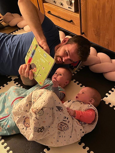 mężczyzna leży na podłodze z dwojgiem niemowląt i czyta im książkę