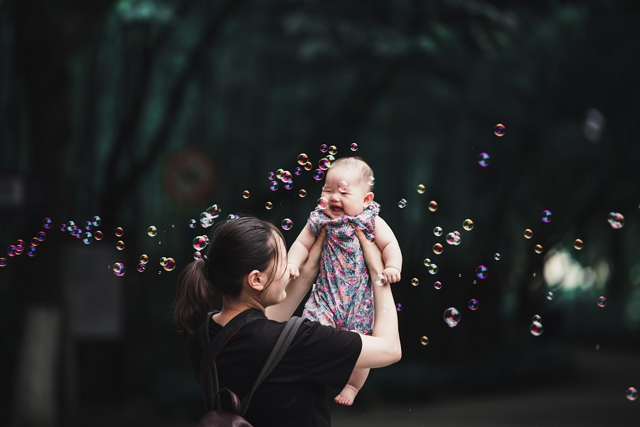 kobieta o azjatyckich rysach podrzuca w górę dziecko; w tle bańki mydlane