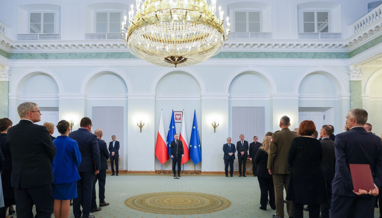 mężczyzna stoi na tle flag Polski i UE; na pierwszym planie dwie grupy ludzi cyłem do obiektywu, MęFot. Kancelaria Prezydenta RP