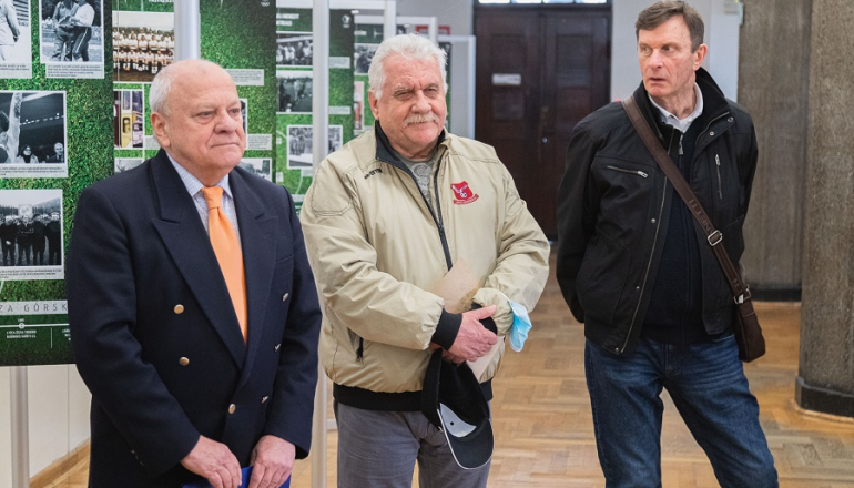 trzech mężczyzn stoi na tle plansz z wystawy o trenerze Kaziemierzu Górskim