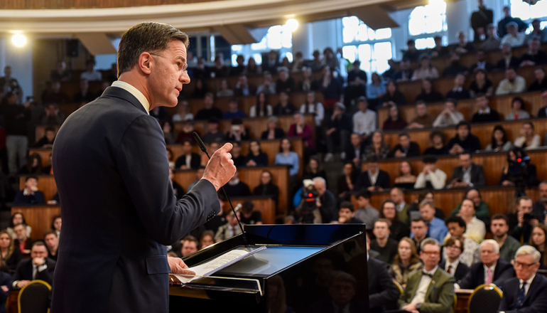 mężczyzna za mównicą przemawiający do pełnej ludzi auli