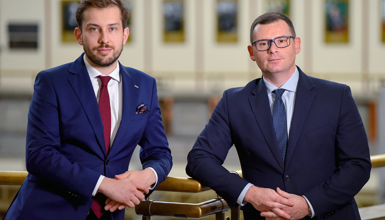 Dwóch mężczyzn w garniturach, stoją bark w bark, patrzą przed siebie
