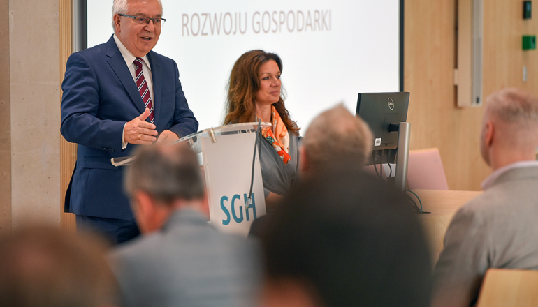 Mężczyzna i kobieta występujący przed słuchaczami, za nimi tablica z tematem: przestrzenne uwarunkowania rozwoju gospodarki