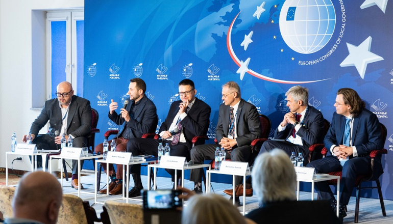sześcioro mężczyzn dyskutuje za stołem na kongresie; w tle plansza przedstawiająca siatkę kuli ziemskiej z flagą UE i gwiazdami wokół siatki symbolizującymi UE
