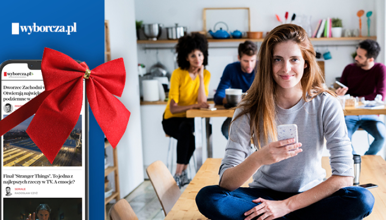 na pierwszym planie kobieta trzymająca komórkę, obok telefon komórkowy z czerwona kokardą
