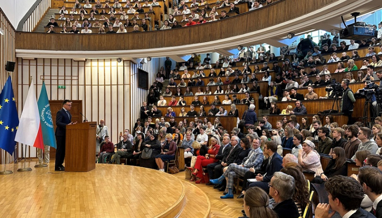 aula pełna studentów i wykładowców; na scenie przemawia mężczyzna