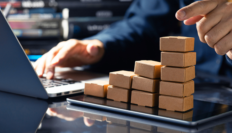 na stole paczki ułożone jedna na drugiej, otwarty laptop i dłoń wskazująca stos paczek