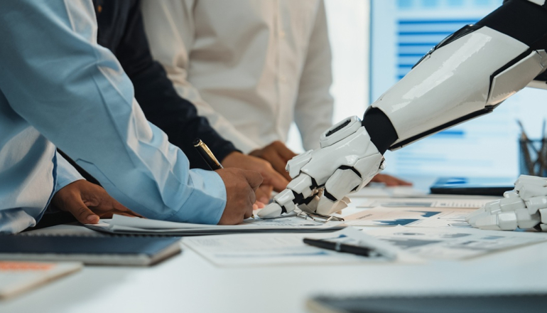 dwie osoby (widać tylko tułowie) dyskutują nad jakimś planem na biurku, pokazując w jedno miejsce palcami; z prawej strony widać także rękę robota