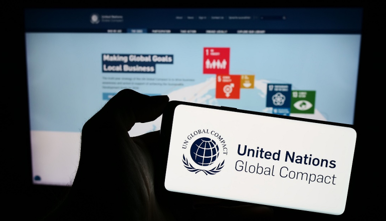 w dłoni trzymany telefon komórkowy, na którym wyświetla sie logo i napis United Nations Global Compact, w tle na dużym ekranie wyswietlana prezentacja