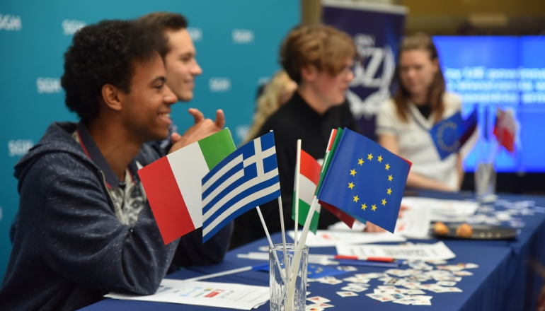kilka osób siedzi za stołem; na pierwszym planie kilka flag, w tym flaga UE
