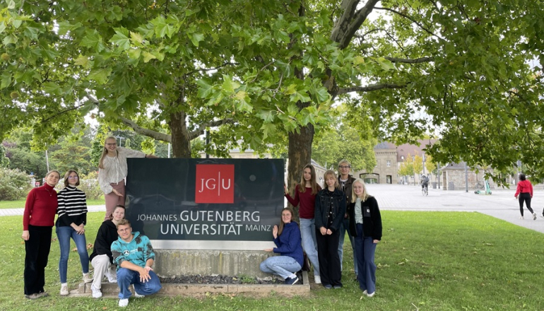 grupa młodych ludzi stoi przy tablicy informacyjnej; w tle drzewa i zieleń