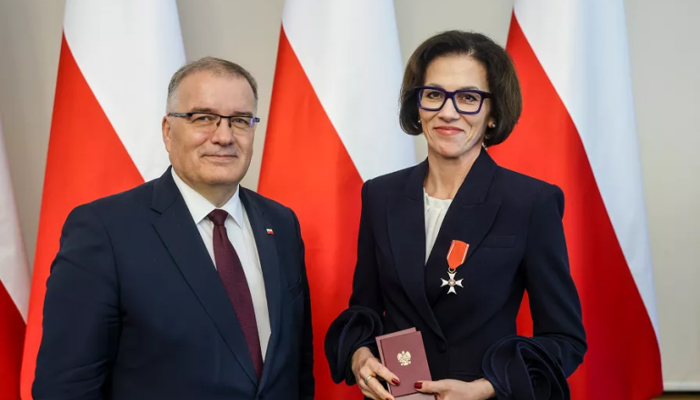 kobieta przyjmuje odznaczenie; zdjęcie na tle trzech polskich flag