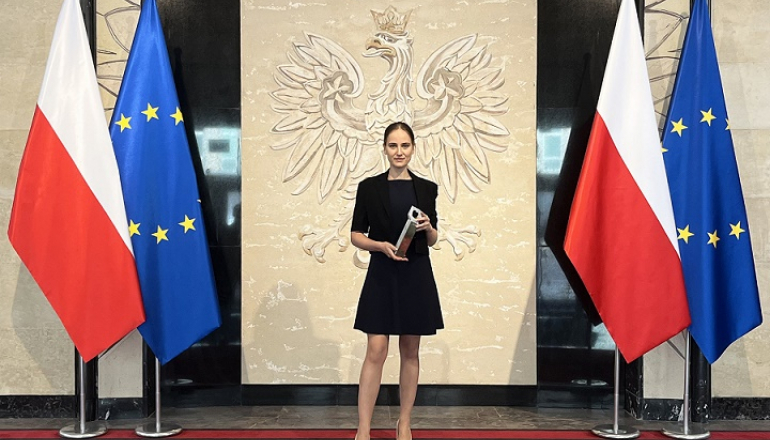 kobieta w czarnej sukience na tle godła RP; po bokach dwie flagi PL i UE