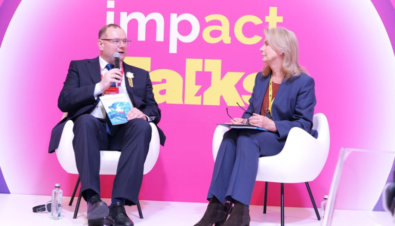 Kobieta i mężczyzna rozmawiają na panelu; w tle różowa ścianka