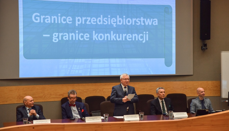 kilku mężczyzn na konferencji naukowej siedzi za stołem prezydialnym, jeden stoi i przemawia