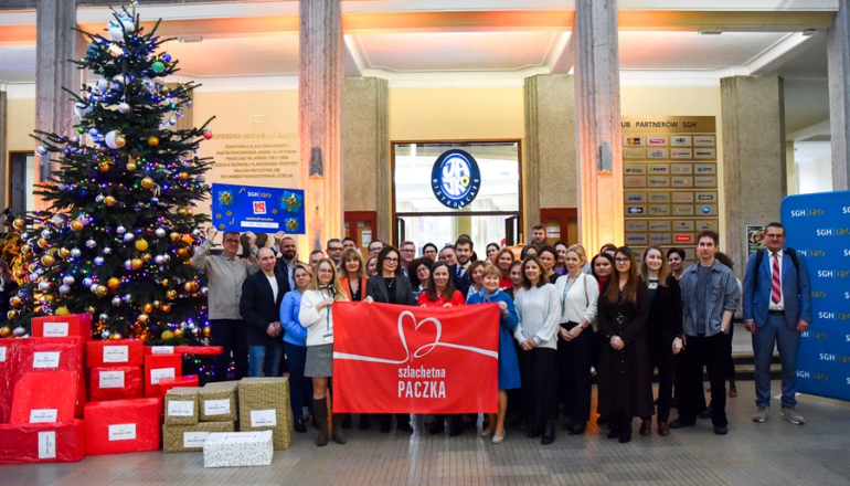 kilkanaście osób pozuje do zdjęcia przy dużej choince i prezentach 