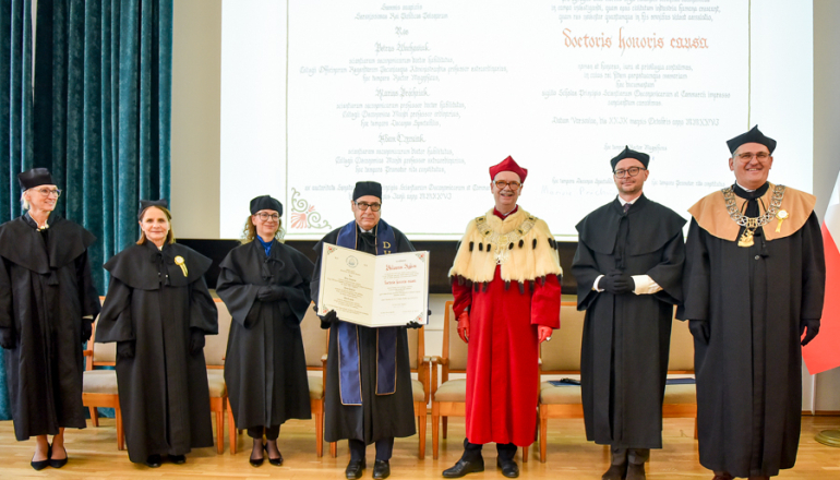 zdjęcie z uroczystości na uczelni - kilka osób w togach stoi na scenie