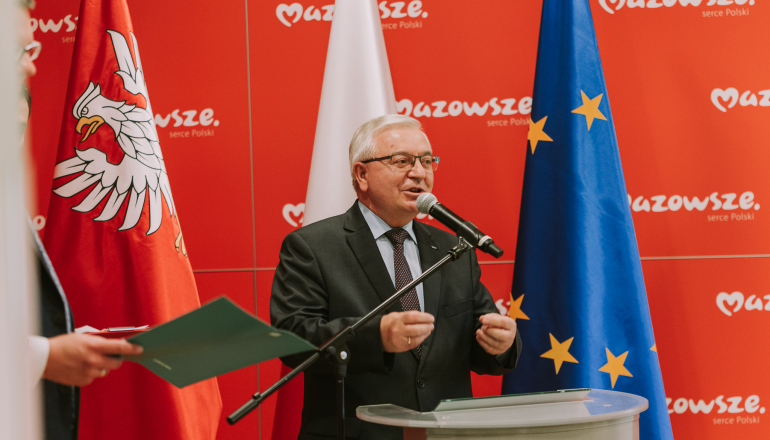 mężczyzna stojący przy mównicy na tle flagi polskiej i unijnej