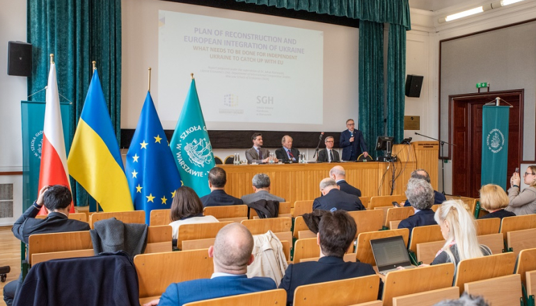 grupa osób siedzi na sali, na scenie kilka osób siedzi za stołem prezydialnym; po lewej stronie sceny flagi Polski, Ukrainy, UE, SGH