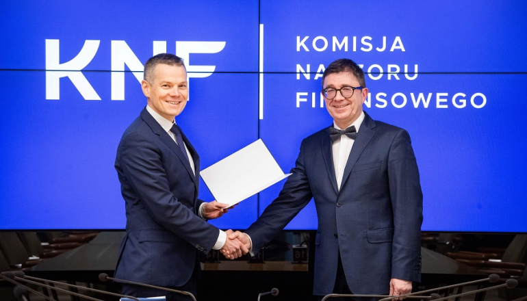 mężczyzna przekazuje dyplom drugiemu mężczyźnie; w tle niebieskie tło z logiem i nazwą KNF