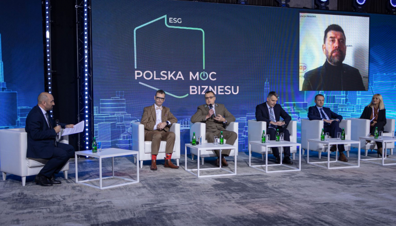 zdjęcie kolorowe. Na krzesłąch postawionych na scenie siedzi pięć osób obok siebie. Obok siedzi osoba, która prowadzi dyskusję