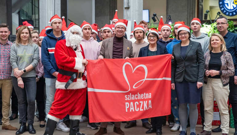 grupa osób w czapkach mikołajowych stoi przed ubraną choinką; częśc z nich w rękach trzyma czerwony transparent z napisem Szlachetna Paczka