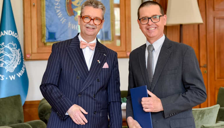 dwóch mężczyzn stoi w gabinecie; jeden z nich trzyma w ręku teczkę z nominacją