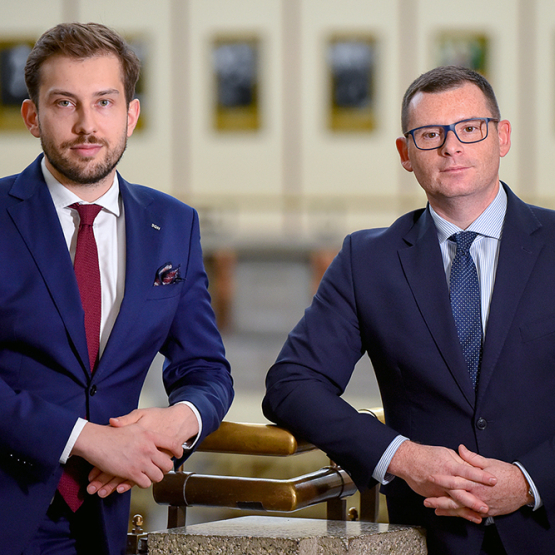 Dwóch mężczyzn w garniturach, stoją bark w bark, patrzą przed siebie