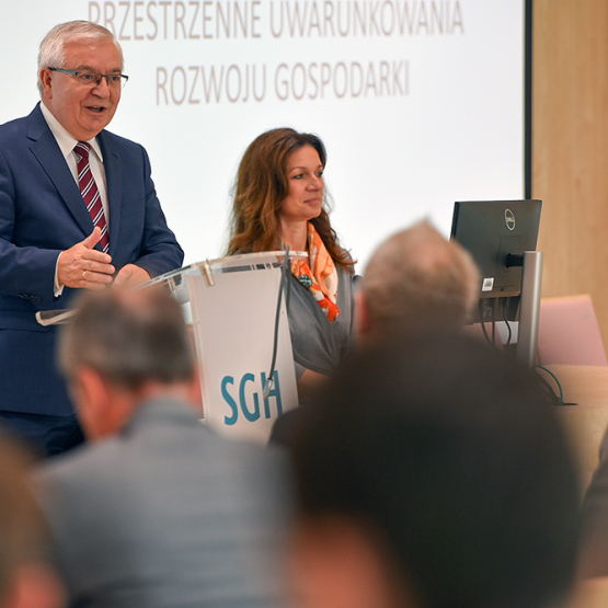 Mężczyzna i kobieta występujący przed słuchaczami, za nimi tablica z tematem: przestrzenne uwarunkowania rozwoju gospodarki