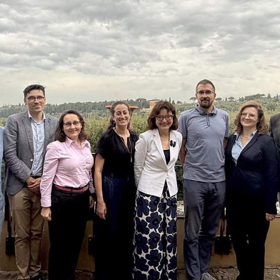 Osiem uśmiechających się osób, stojących na tle toskańskiego krajobrazu 