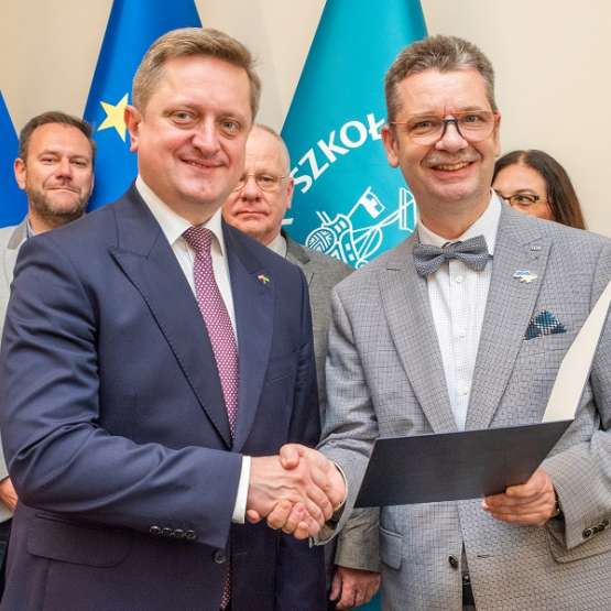 dwóch mężczyzn podaje sobie dłoni po przekazaniu dokumentów; za nimi w tle grono osób