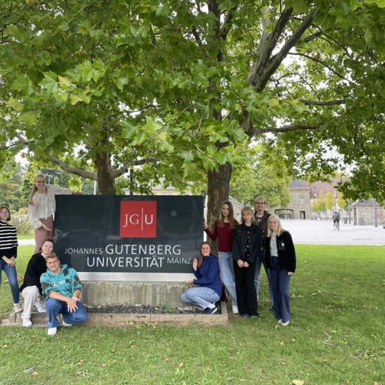 grupa młodych ludzi stoi przy tablicy informacyjnej; w tle drzewa i zieleń