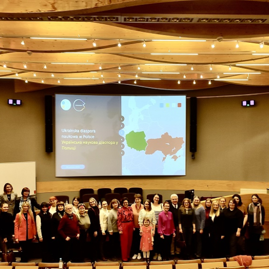 grupa osób stoi w jednej z sal wykładowych; za nimi ekran informujacy o spotkaniu Ukraińskiej diaspory naukowej w Polsce; napis po polsku i ukraińsku