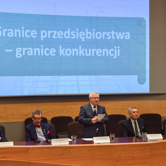 kilku mężczyzn na konferencji naukowej siedzi za stołem prezydialnym, jeden stoi i przemawia