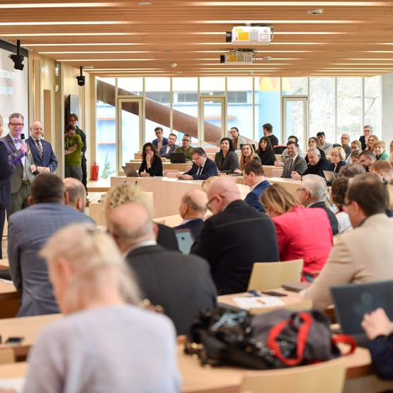 trzech mężczyzn stoi i przemawia w sali konferencyjnej wypełnionej ludźmi