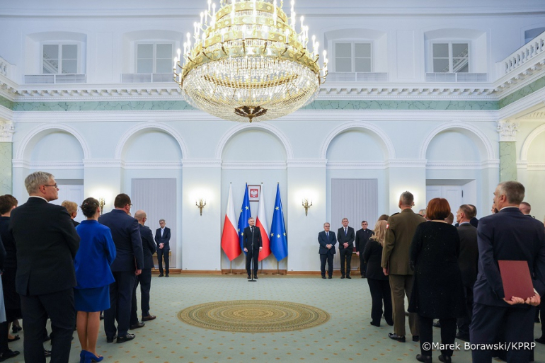 mężczyzna stoi na tle flag Polski i UE; na pierwszym planie dwie grupy ludzi cyłem do obiektywu, MęFot. Kancelaria Prezydenta RP