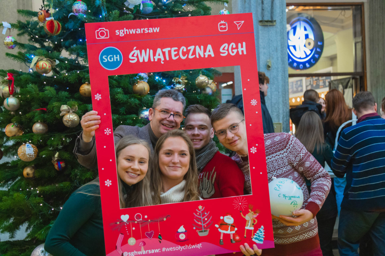 grupa osób stoi przed ubraną choinką; w rękach trzymają ramkę z napisem "Świąteczna SGH"