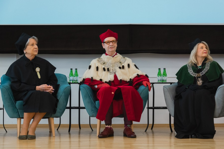 troje osób ubranych w togi siedzi, słuchając wykładu