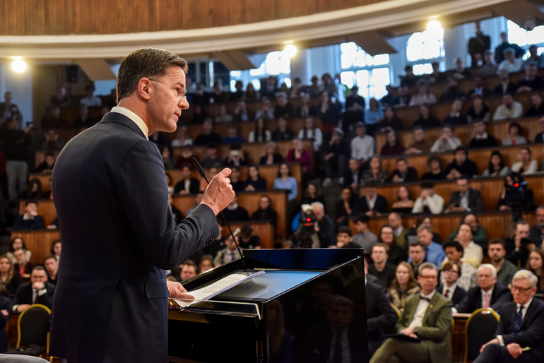 mężczyzna za mównicą przemawiający do pełnej ludzi auli