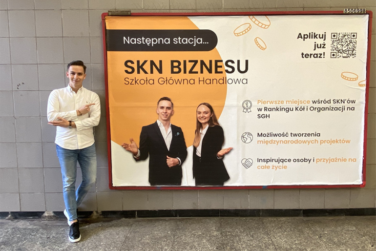 na zdjęciu mężczyzna wskazujący bilbord na ścianie z napisem SKN Biznesu Szkoła Główna Handlowa