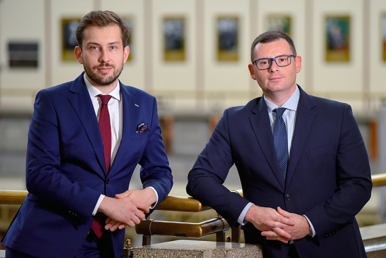 Dwóch mężczyzn w garniturach, stoją bark w bark, patrzą przed siebie