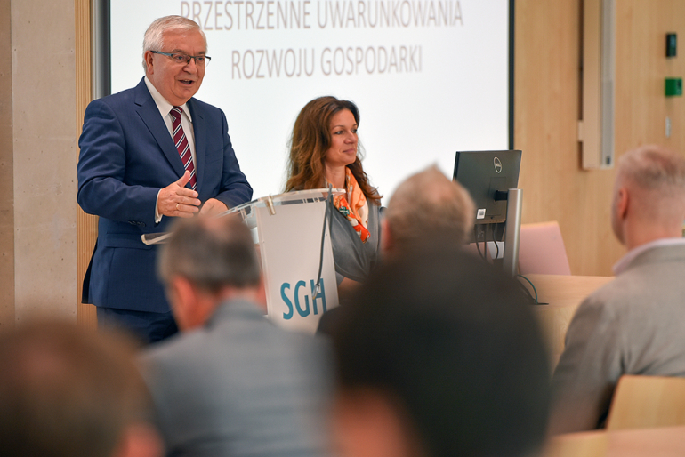 Mężczyzna i kobieta występujący przed słuchaczami, za nimi tablica z tematem: przestrzenne uwarunkowania rozwoju gospodarki