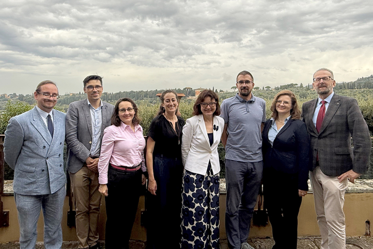 Osiem uśmiechających się osób, stojących na tle toskańskiego krajobrazu 