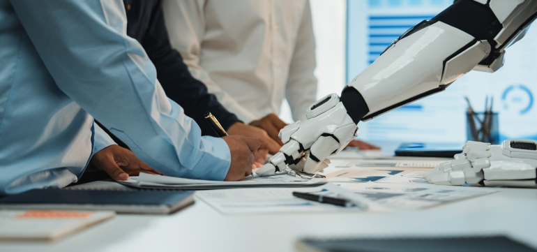 dwie osoby (widać tylko tułowie) dyskutują nad jakimś planem na biurku, pokazując w jedno miejsce palcami; z prawej strony widać także rękę robota