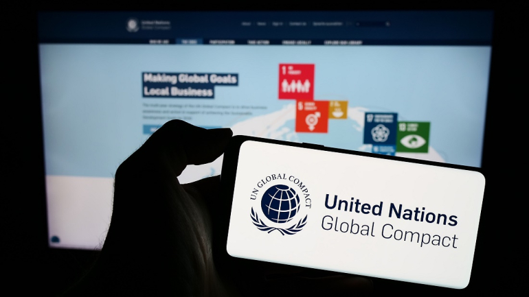 w dłoni trzymany telefon komórkowy, na którym wyświetla sie logo i napis United Nations Global Compact, w tle na dużym ekranie wyswietlana prezentacja