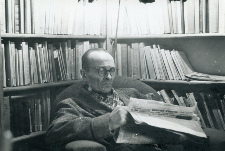 czarno-białe zdjęcie mężczyzny siedzącego w gabinecie pełnym książek i czytającego gazetę