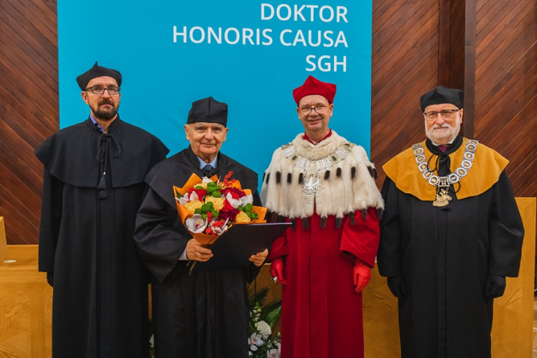 czterech mężczyzn ubranych w togi, w tym jeden w todze rektorskiej, stoją na katedrze w jednej z auli uczelni; z tyłu napis doktor honoris causa SGH