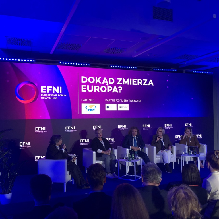 grupa osób siedzi na sali podczas panelu, na scenie kilku prelegentów