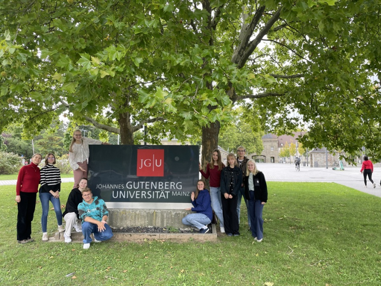 grupa młodych ludzi stoi przy tablicy informacyjnej; w tle drzewa i zieleń